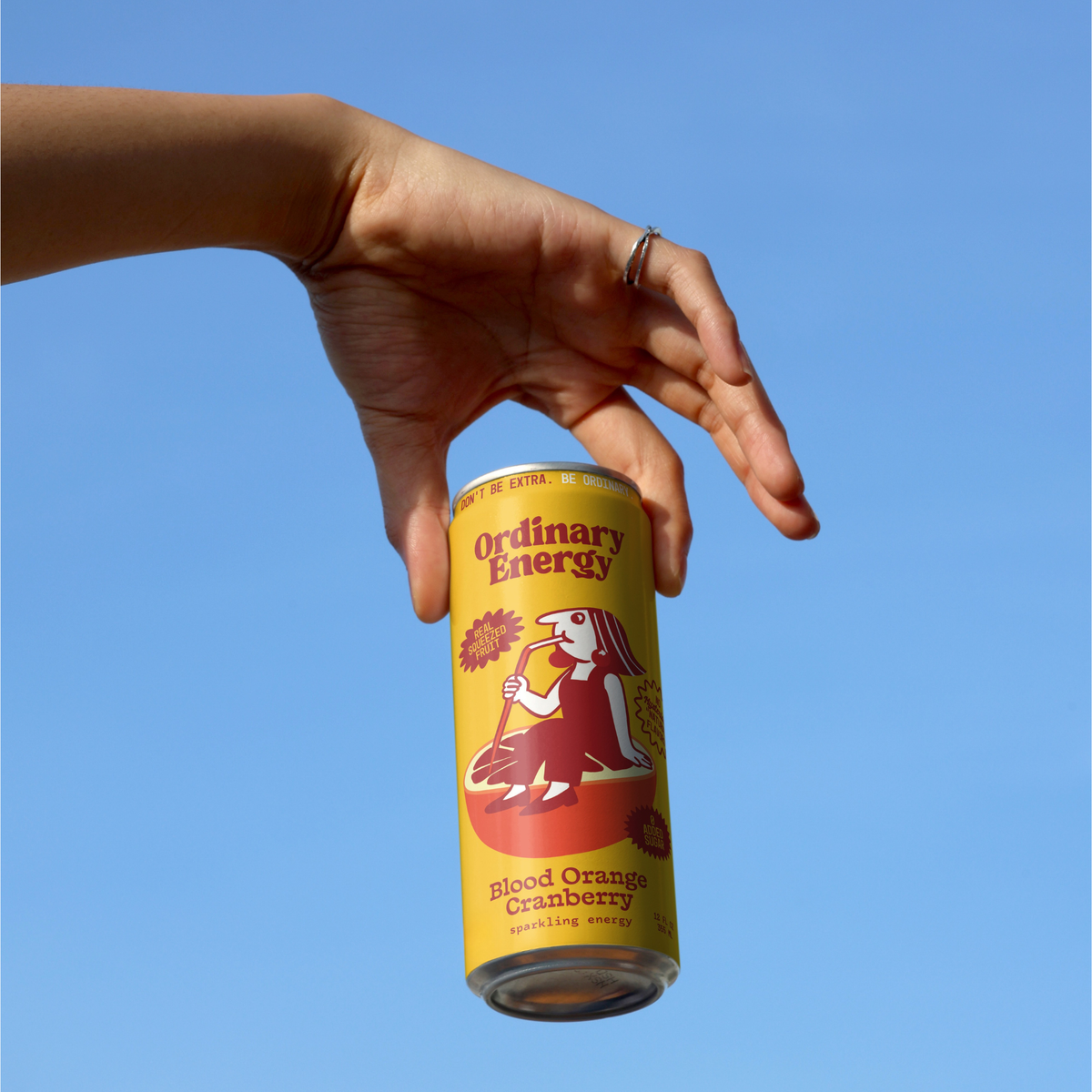 Hand holding a can of Ordinary Energy Blood Orange Cranberry against a blue sky